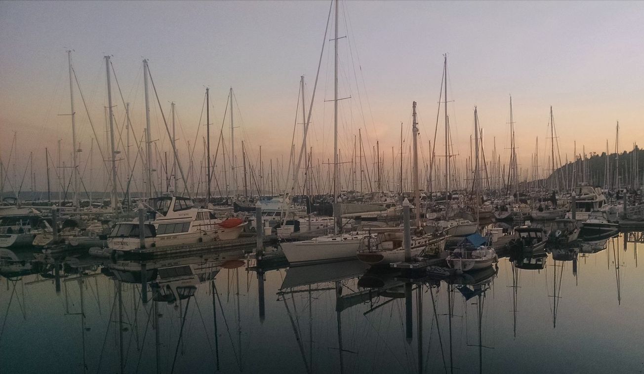Dozens of boats at Shilshole Marina. The glow of a sunrise coats the clouds in the sky. The masts of the sailboats are refl