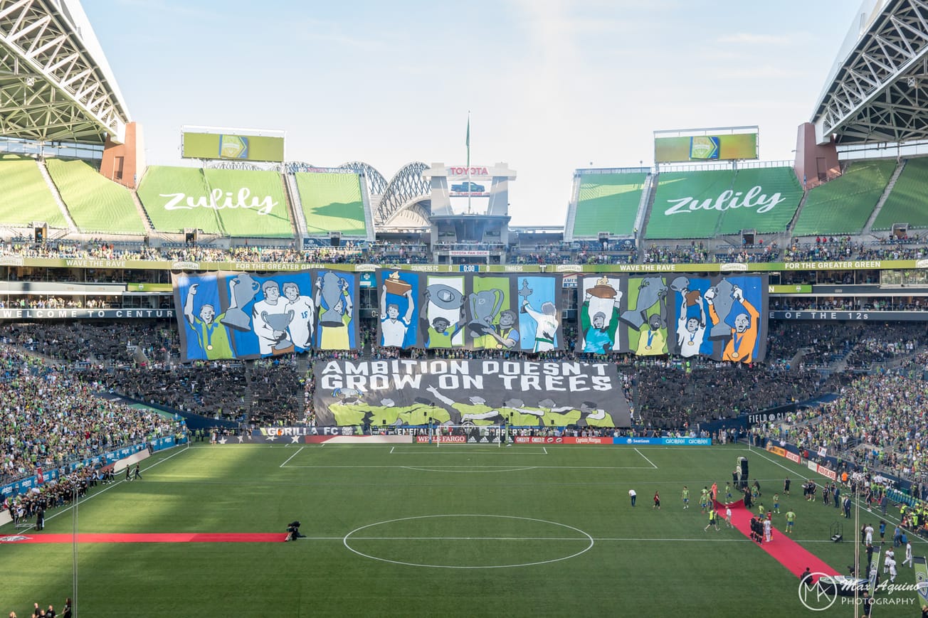 Photo of an ECS tifo which says "Ambition Doesn't Grow On Trees" and shows Sounders holding many trophies.