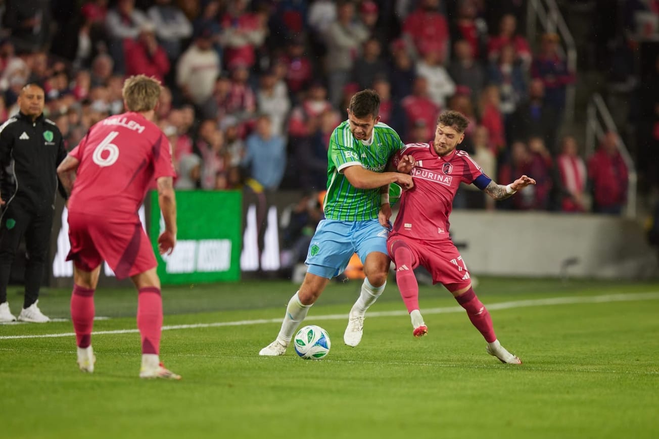 Jackson Ragen, wearing a green jersey and blue shorts, fights for the ball with a player in red.