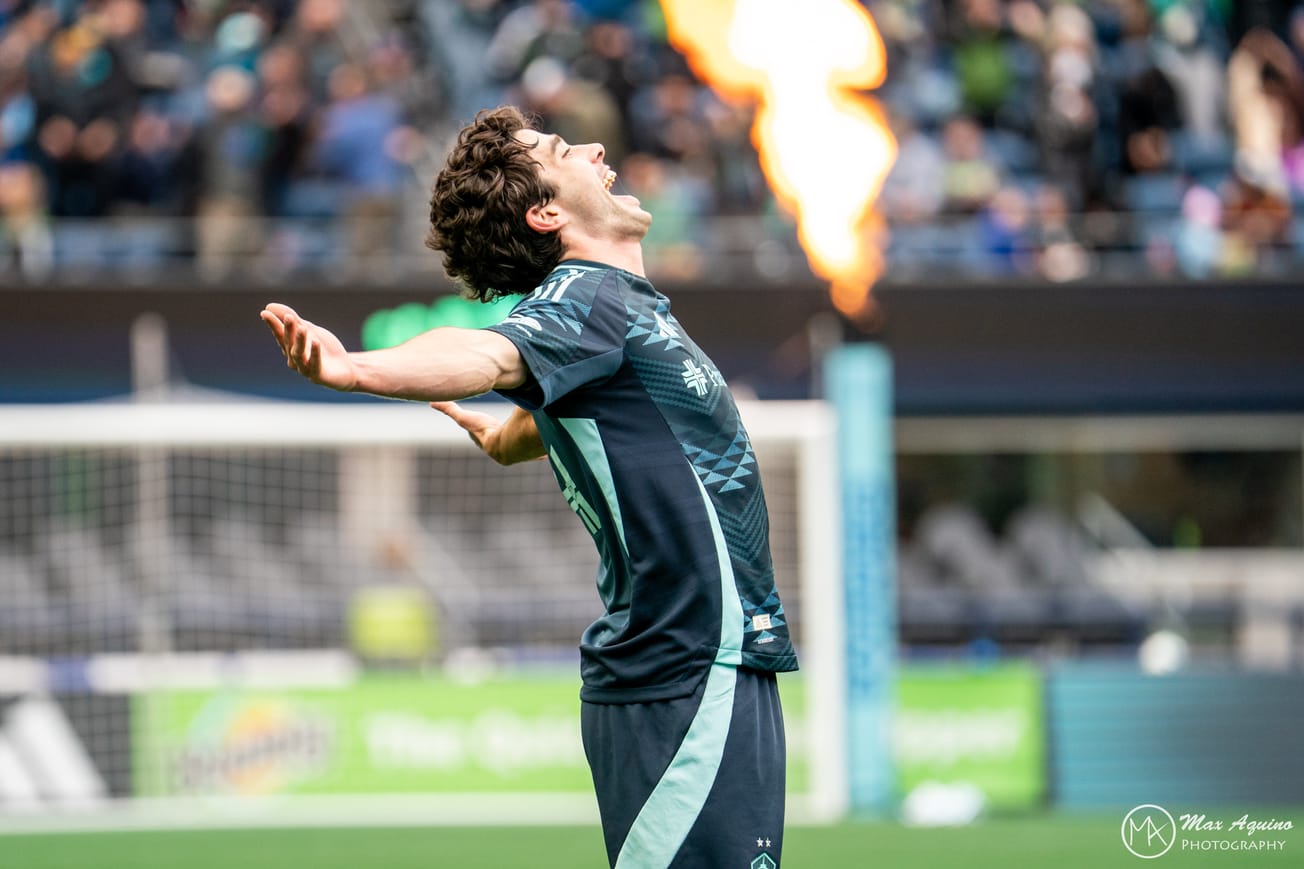 Paul Rothrock celebrates his goal with his arms spread wide and a flamethrower in the background.