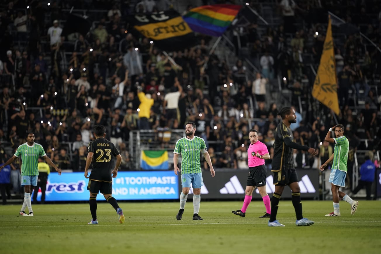 João Paulo in a green Sounders shirt with blue shorts stands in front of the flag-wavin crowd