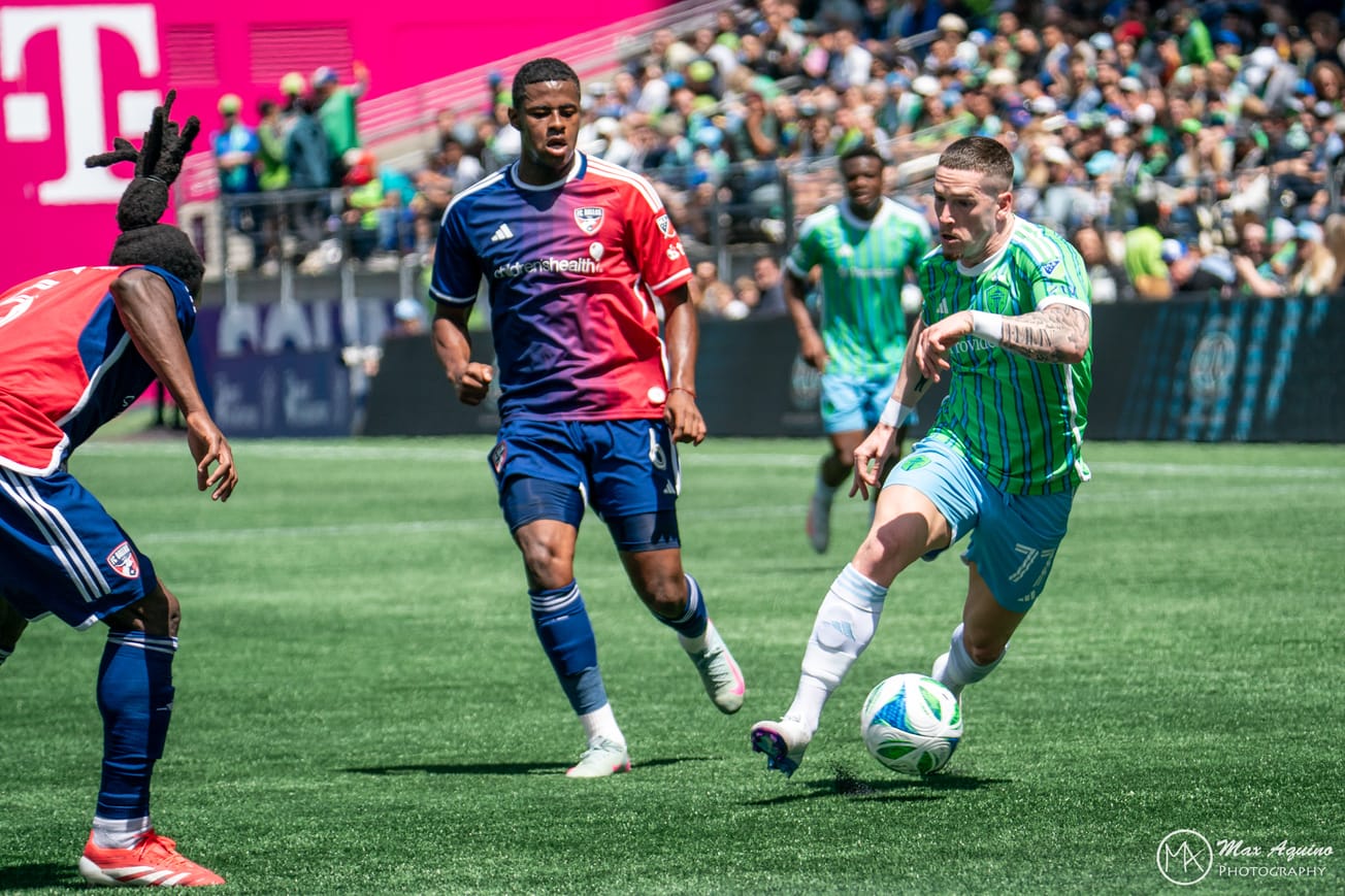 Ryan Kent wearing a green Sounders jersey and light blue shorts dribbles at two Dallas defenders in red and blue.