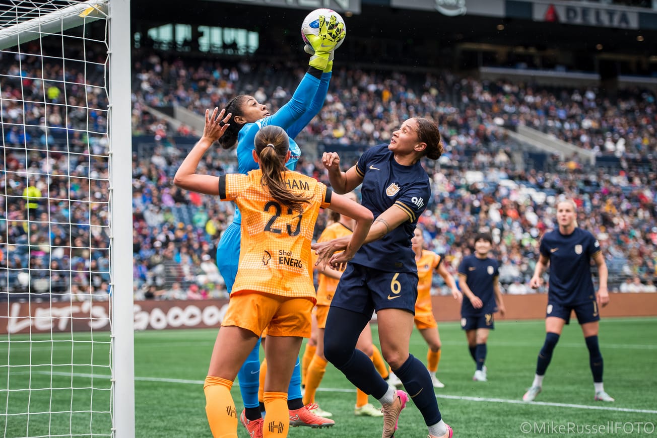 Houston Dash goalkeeper Abby Smith leaps to claim a ball over Reign forward Lynn Biyendolo and several Dash teammates in a May 2025 match.