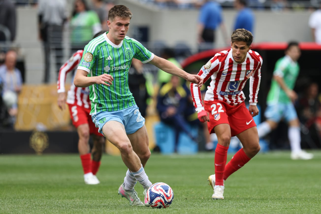 Reed Baker-Whiting wearing a green Sounders jersey with light blue shorts dribbles a red, white and blue ball ahead of Atletico Madrid players in red and white.