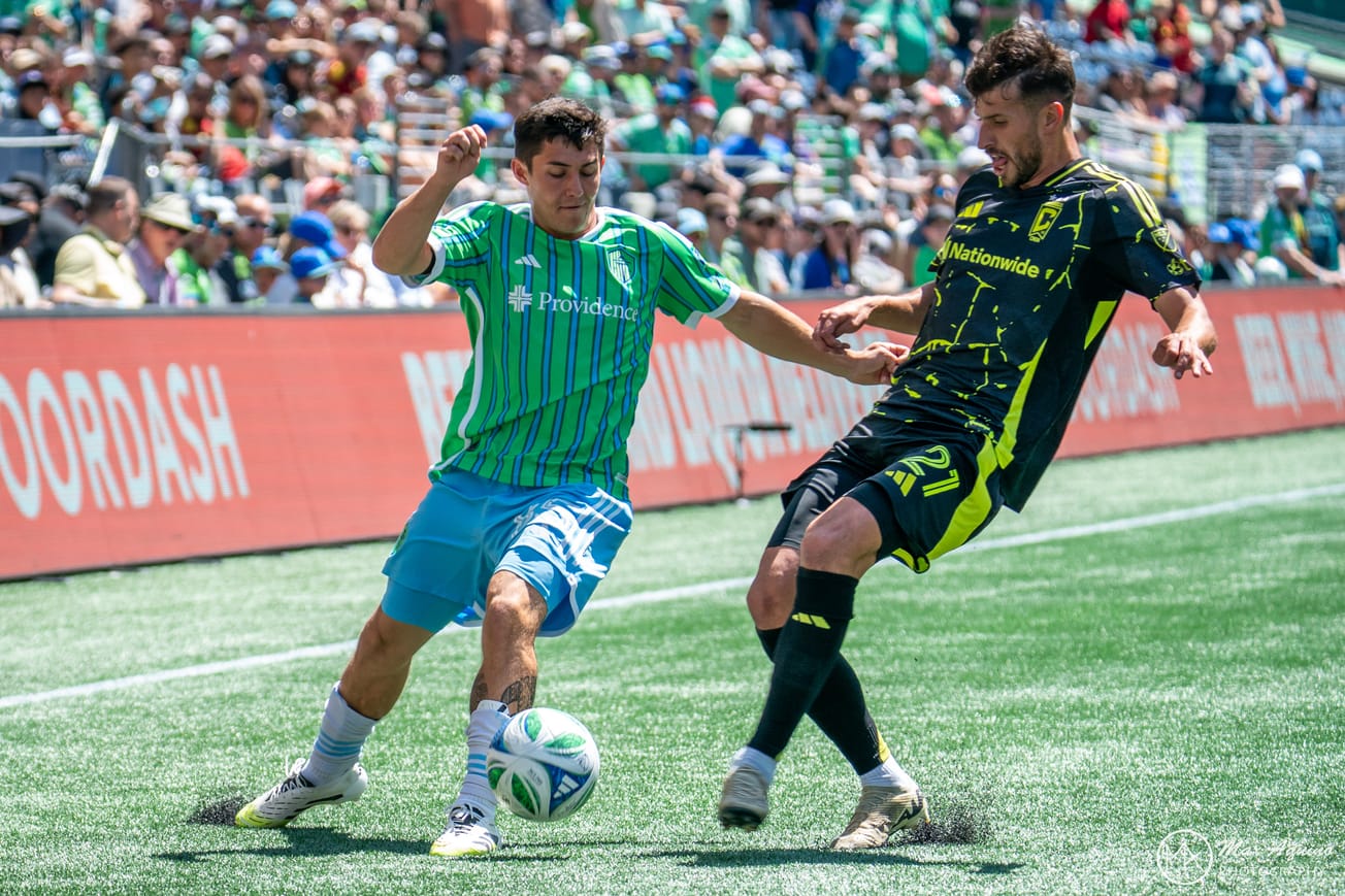 Kalani Kossa-Rienzi in light blue shorts and a striped green Sounders jersey challenges for the ball.