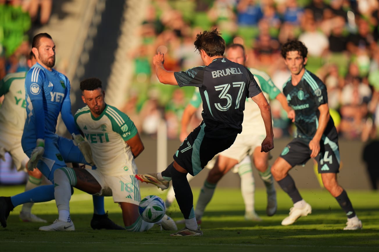 Snyder Brunell, in a dark blue Sounders kit, shoots a ball at the Austin FC goalkeeper