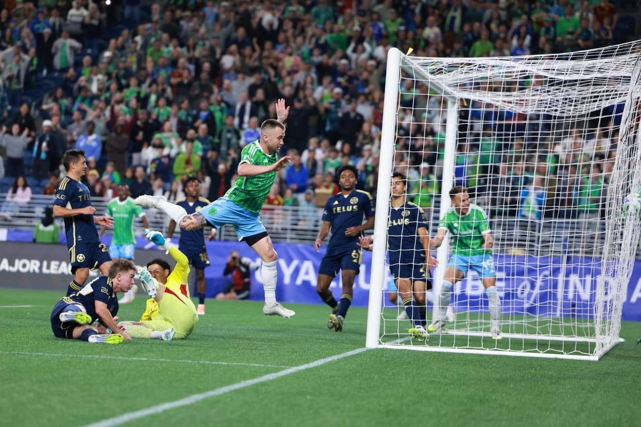 Albert Rusnak wearing a green Sounders Hirt and blue shorts leaps over the goalkeeper after scoring his goal.
