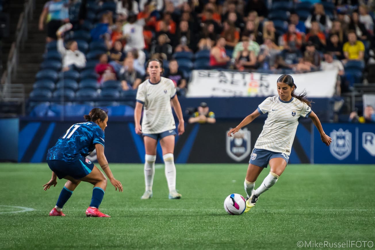 Seattle Reign midfielder Sam Meza dribbles the ball against a San Diego Wave player as teammate Phoebe McClernon looks on in an August 2025 match at Lumen Field.