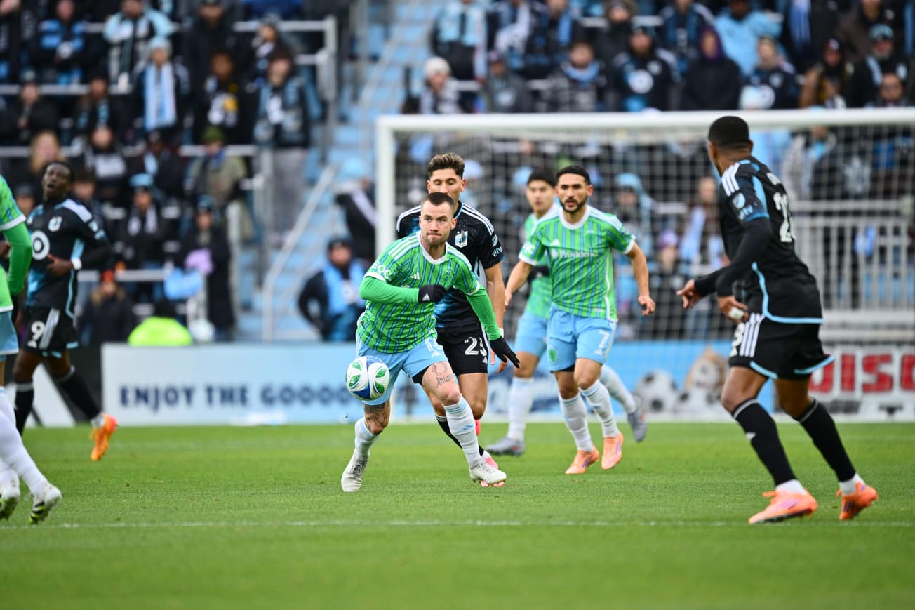 Albert Rusnak in a green Sounders jersey and light blue shorts has the ball surrounded by Minnesota players.