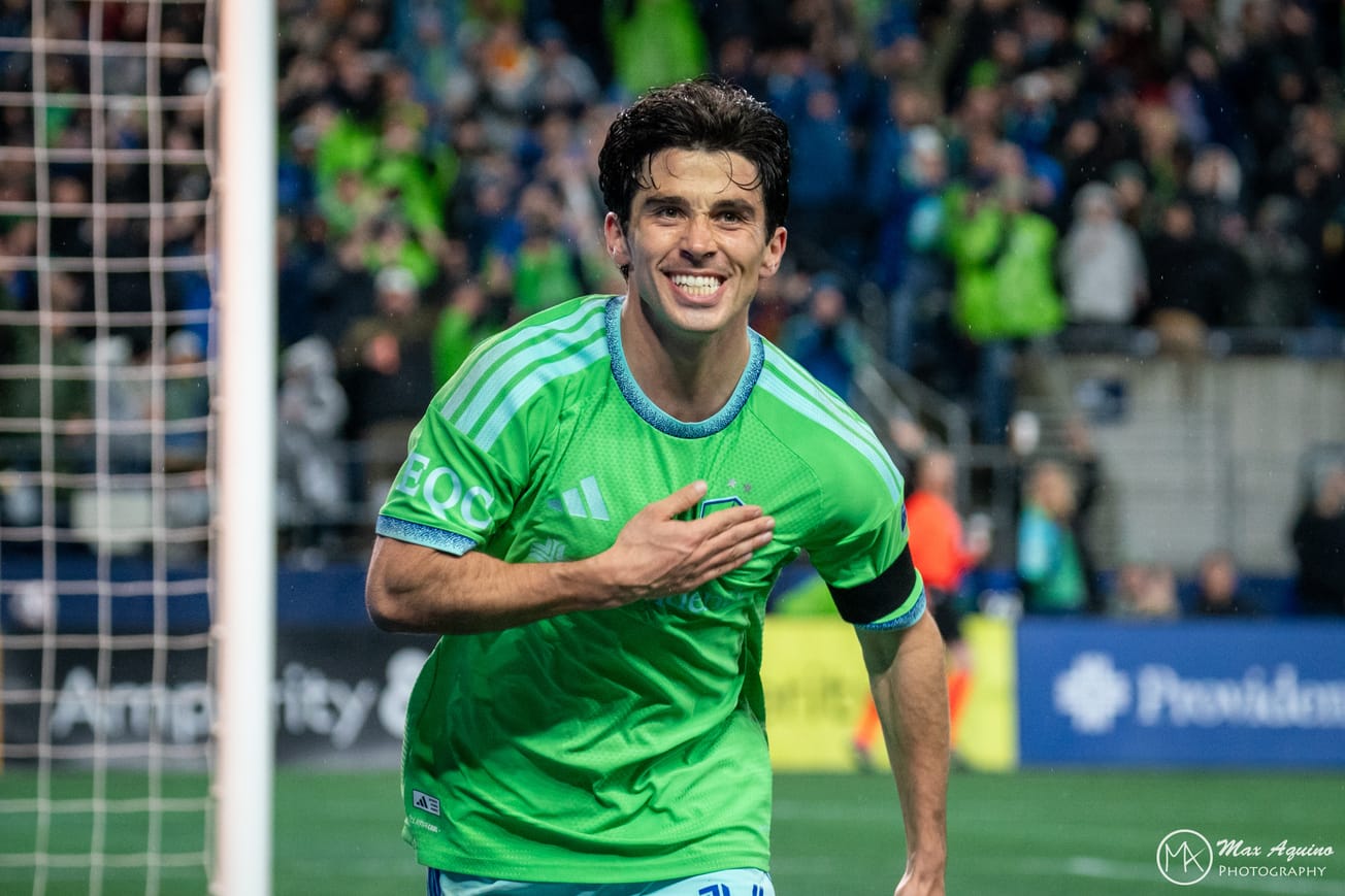 Paul Rothrock wearing a green Sounders jersey smiles and pats the Sounders badge after scoring a goal.