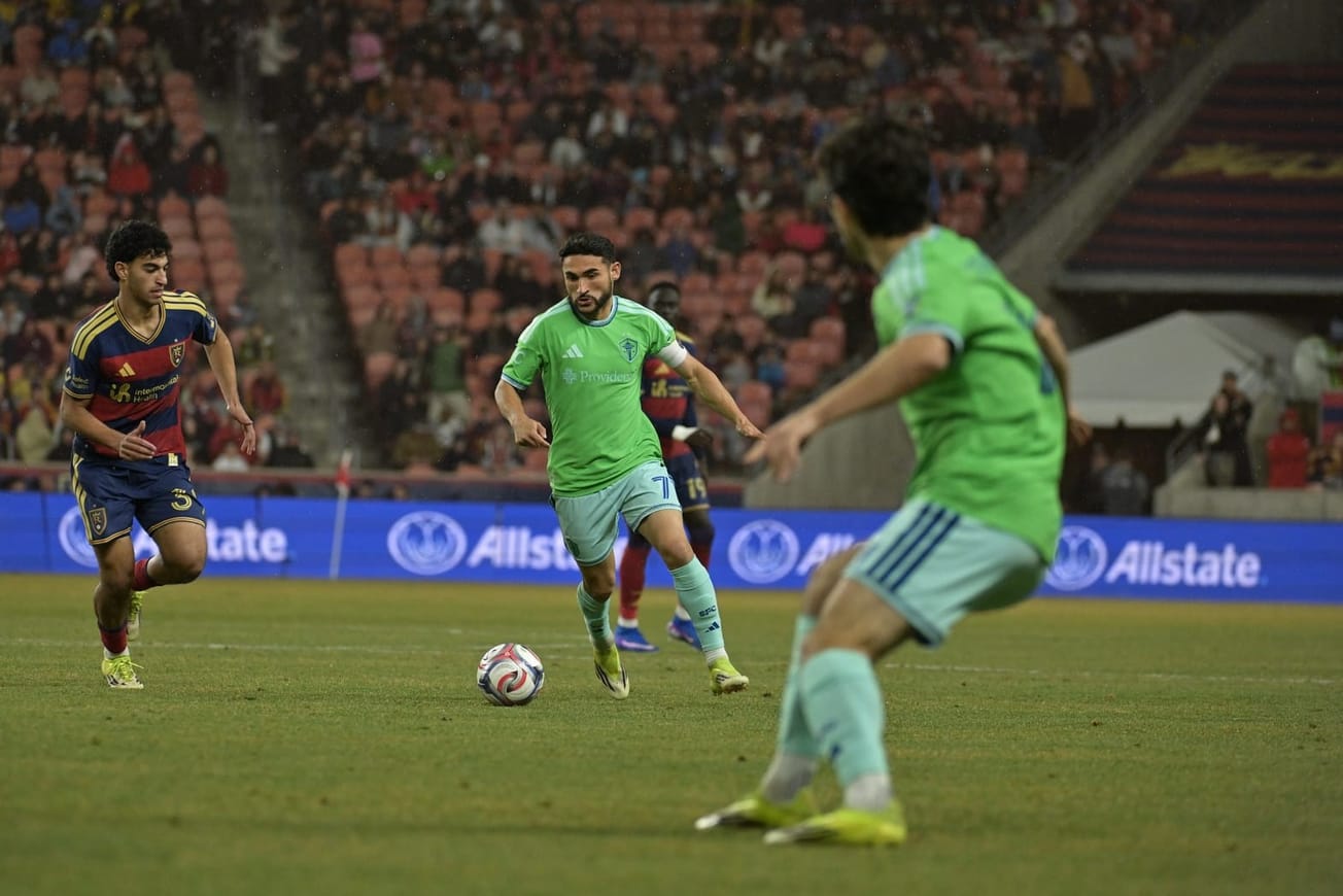 Cristian Roldan wearing a green Sounders jersey and light blue shorts dribbles with an RSL player next to him and Paul Rothrock in the foreground.