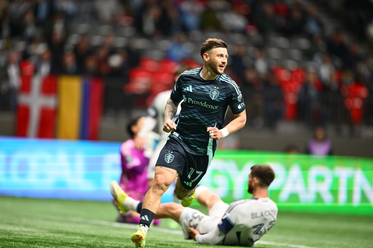 Paul Arriola is wearing a blue Sounders kit and is running to celebrate a goal while Whitecaps players lay on the ground in the background.