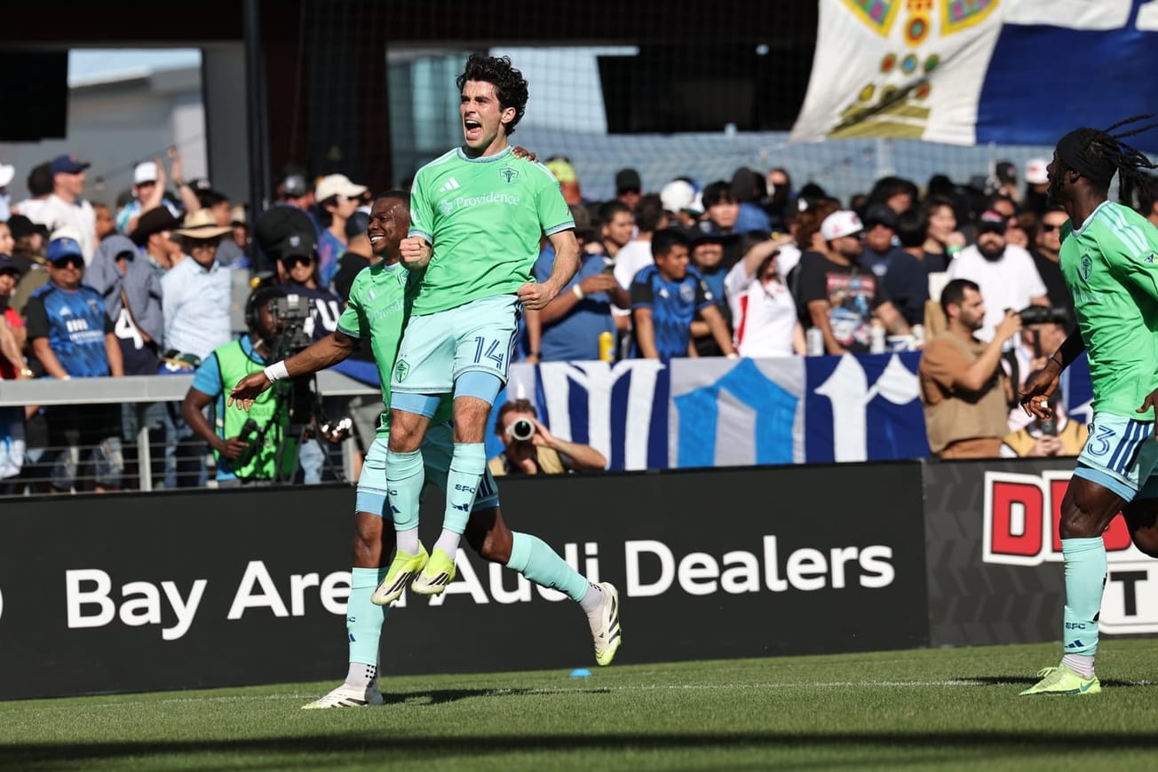 Paul Rothrock, wearing a green Sounders jersey and light blue shorts and socks, leaps in the air celebrating a goal with Nouhou and Georgi Minoungou running behind him.